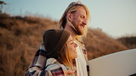 Close-up-shot-of-a-happy-blond-guy-with-a-beard-in-a-plaid-shirt-hugs-his-blonde-girlfriend-in-a-hat-in-a-plaid-shirt-and-holds-a-surfboard-near-the-strawberry-seashore-at-Sunrise-in-the-morning-in-summer