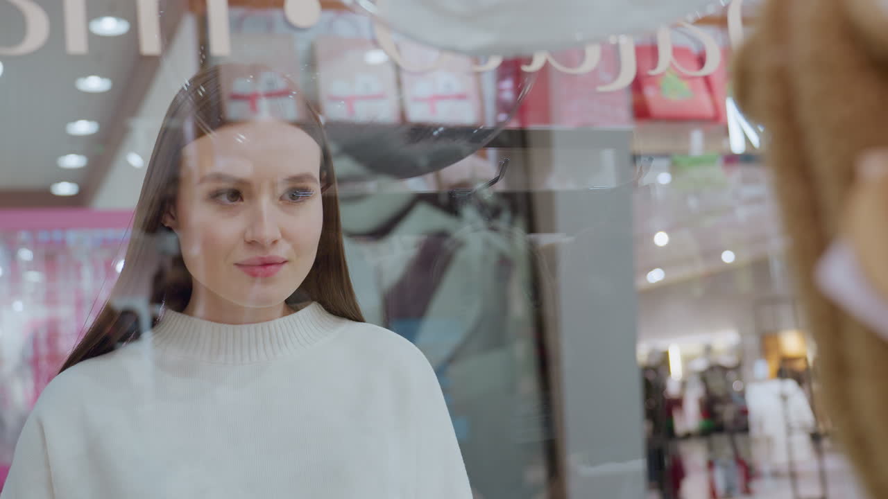 Young lady looking at something in the background of a retail store, her reflection is clearly visible on the glass as she admires the display, bright and modern mall setting