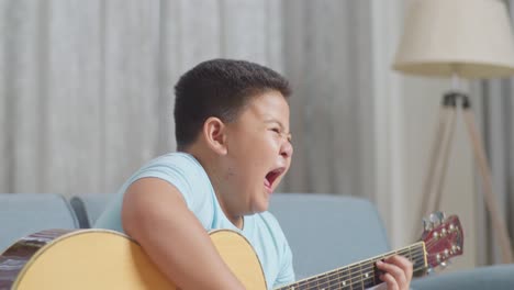 close up of talented asian kid with expressive face enjoys playing the guitar on sofa at home