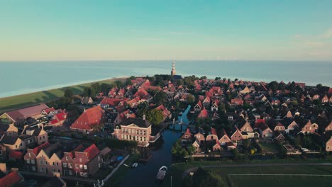 Coastal-town-at-sunset-with-lighthouse,-aerial-view-of-red-rooftops,-calm-evening