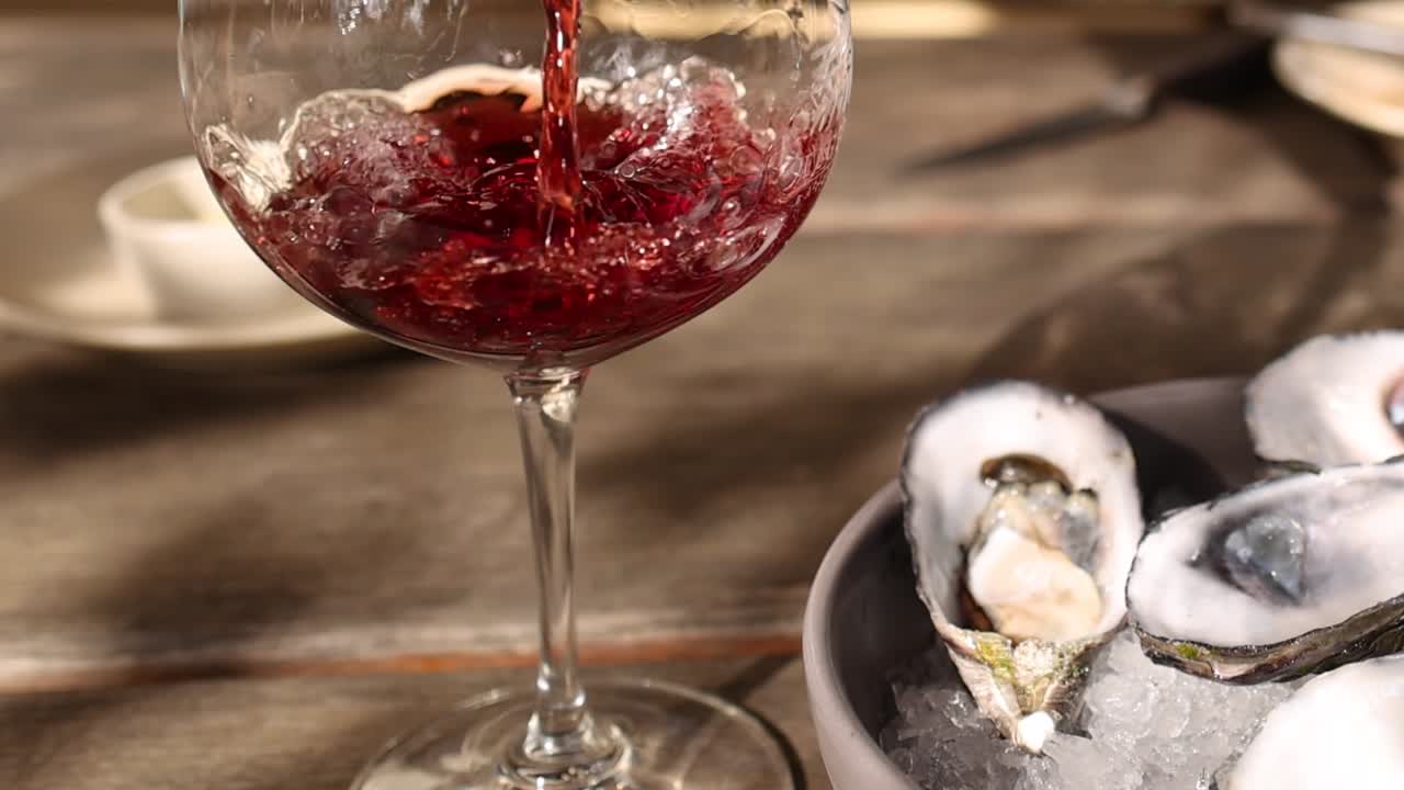 Vino rosso viene versato in un bicchiere di cristallo accanto a una ciotola di ostriche fresche.