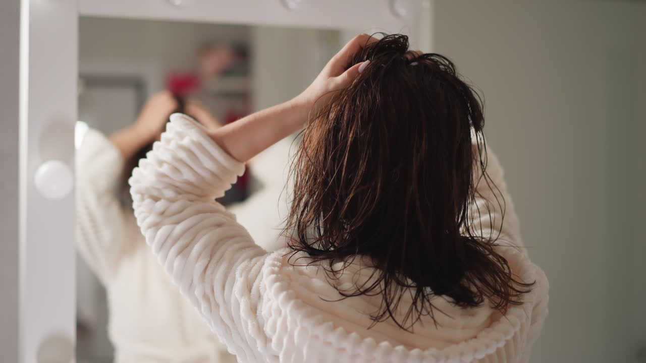 은 옷을 입은 여성이 화장실에서 은 날아다니는 머리카락을 회전시니다. 여성은 거울에서 흐릿한 반사와 함께 화장실에 머리카락를 조정하는 손을 움직입니다.