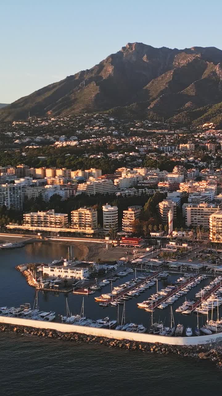 Ripresa aerea verticale con drone del porto di Marbella al tramonto, yacht di lusso sottostanti con la montagna La Concha della Sierra Blanca che si erge sullo sfondo, luce mediterranea incandescente che cattura l'iconica eleganza costiera