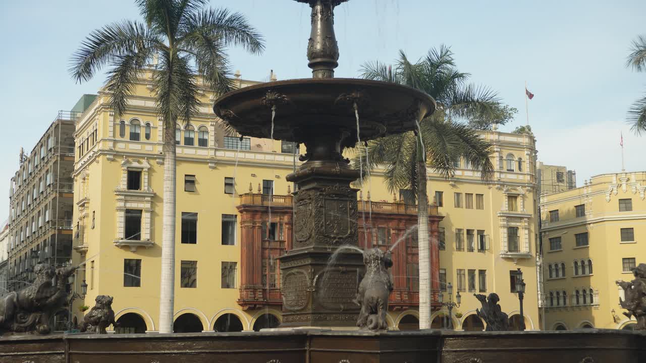 A fountain with water running. Palm trees and yellow building is in the background, with the historic spanish colonial era wooden balcony. Located in the "Plaza Mayor" in the center of Lima, Peru