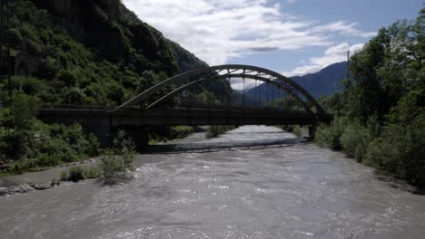 river after a flood flows under a bridge where cars and trucks driving in switzerland in front of big mountains and forest on a sunny day