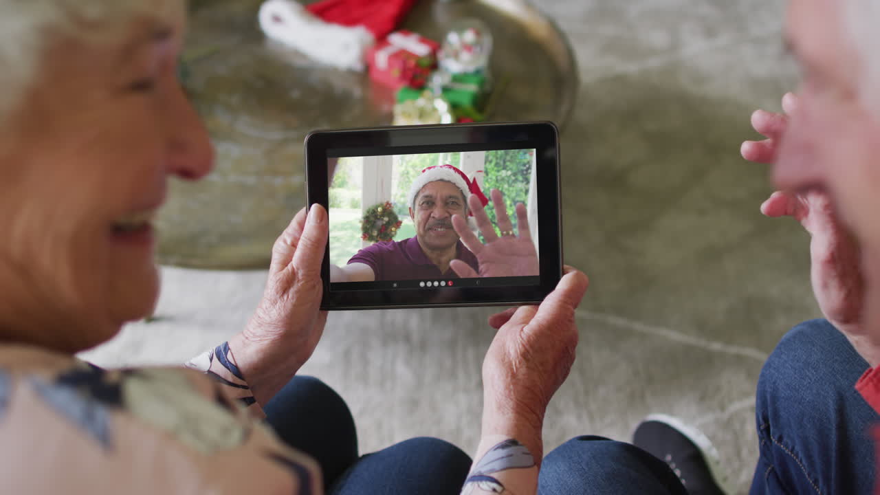 una pareja caucásica mayor sonriente usando una tableta para una videollamada de navidad con un hombre en la pantalla
