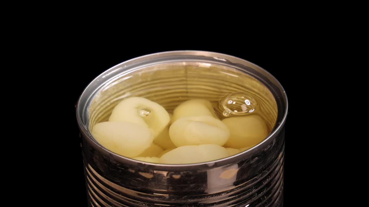 Chopsticks lift and drop a piece of canned longan fruit back into syrup, shot in close-up with bright, even lighting against a black background