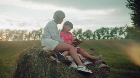 heartwarming side view of a mother embracing her daughter while seated on hay foliage at dusk, reading together, with a hat nearby, surrounded by an open countryside field