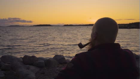 a man is sitting on a rocky beach looking out at the ocean with pipe in his mouth the sky is golden sunset which casts an orange hue across the horizon creating a silhouette of the person calm smoking