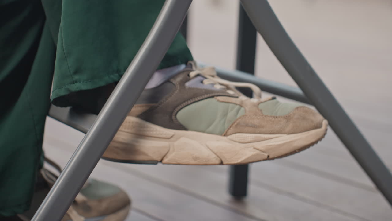 scarpa da ginnastica sotto una sedia, piede seduto in posizione rilassata. Primo piano di scarpa da ginnastica verde consumata e pantaloni verdi con risvolto appoggiati sulla gamba di una sedia di metallo, luce naturale soffusa su un ponte di legno, pelle scamosciata graffiata e suola sporca, lacci