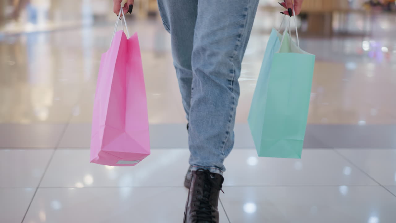 vista de la pierna de una mujer que lleva bolsas de compras rosadas y de menta caminando por el piso pulido del centro comercial que refleja la luz, el fondo borroso con compradores indistintos y pantallas minoristas