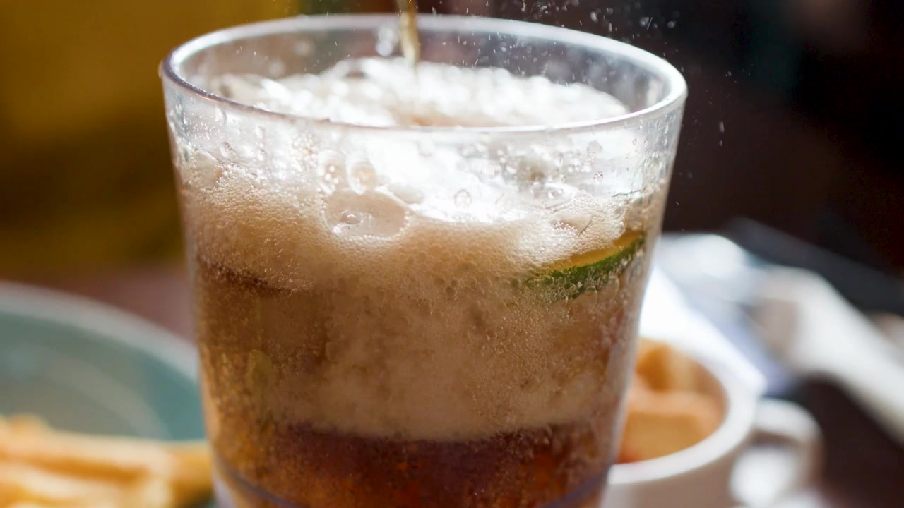 Nahaufnahme von sprudelnder Limonade, die in ein Glas mit Eiswürfeln und Limettenscheiben gegossen wird.