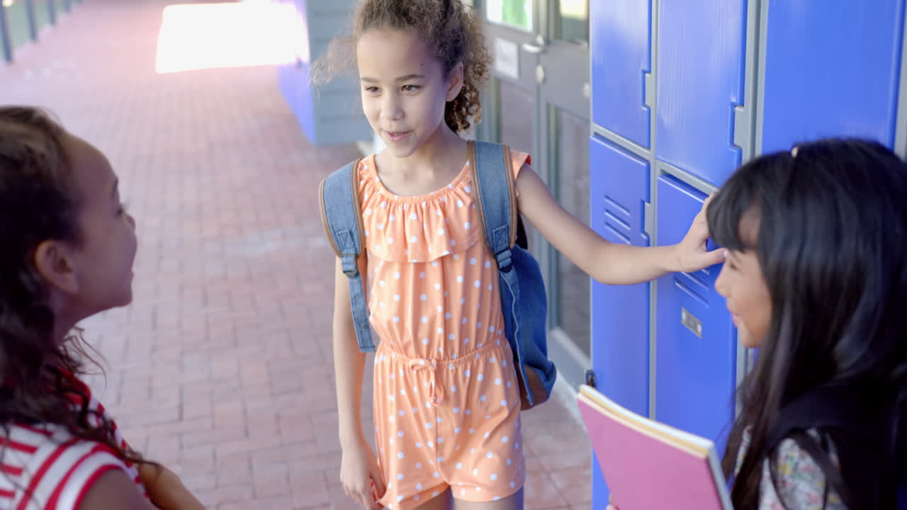 Biracial girls are chatting by school lockers, one wearing an orange polka-dot dress
