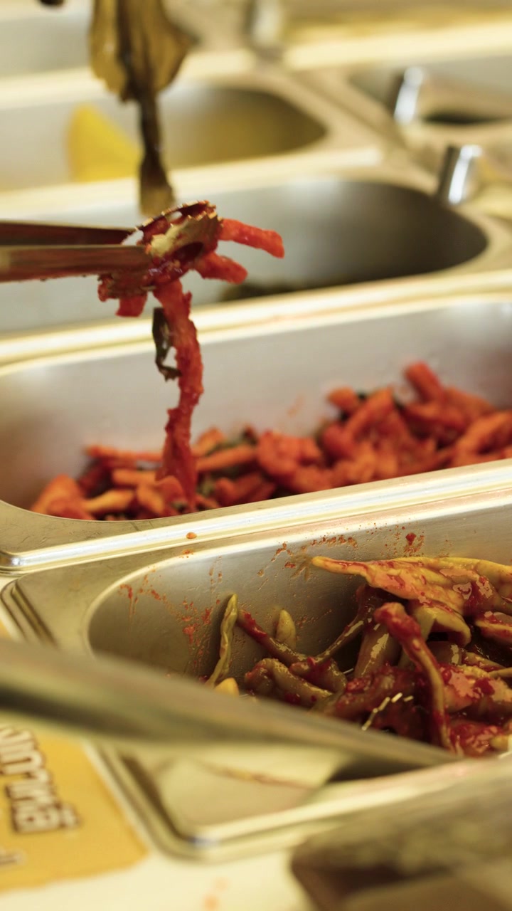Nærbillede af hånd, der bruger metaltang til at vælge krydret kimchi fra restaurantbakker i rustfrit stål