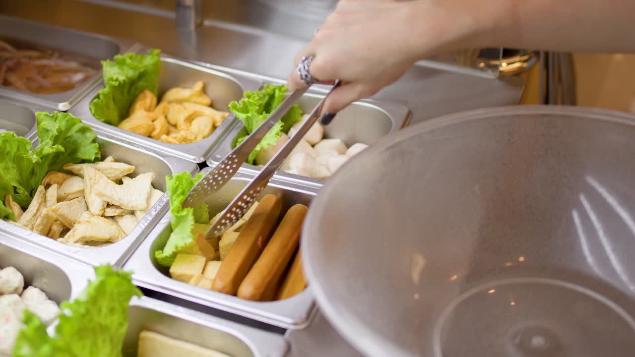 Hand uses tongs to pick raw hotpot ingredients from buffet trays into large bowl