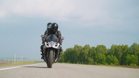 Biker-with-woman-rides-motorcycle-along-road-at-countryside