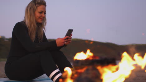 Frau-Sitzt-Auf-Einem-Surfbrett-Am-Lagerfeuer-Am-Strand-Und-Benutzt-Ihr-Mobiltelefon,-Während-Die-Sonne-Hinter-Ihr-Untergeht