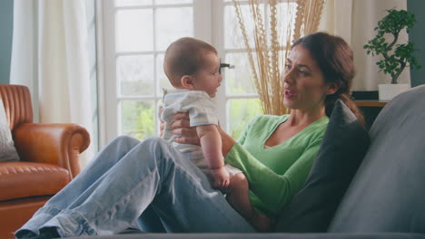 Loving-Mother-Playing-With-Baby-Son-Sitting-On-Sofa-At-Home