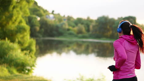 morena con cabello largo en auriculares corre a lo largo del río en el parque por la mañana al amanecer en el verano en una chaqueta rosa y pantalones negros