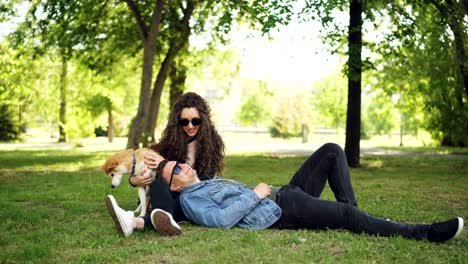 handsome young man is lying on the grass in the park while his girlfriend is talking to him and petting cute dog, couple is laughing and enjoying weekend.