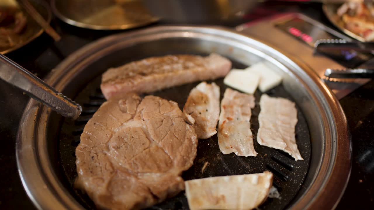 Tongs flip pork slices on tabletop grill, warm lighting, shallow depth, close-up overhead view
