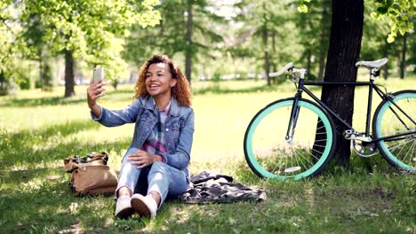una bonita chica afroamericana está haciendo una llamada de video con un teléfono inteligente, está mirando la pantalla, hablando y agitando la mano. es visible un hermoso parque con hierba y árboles.