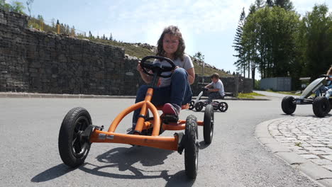 Foto-De-Dolly-Fhd-De-Una-Niña-Pequeña-En-Bicicleta-En-Un-Cuatriciclo-De-Pedal-Naranja-En-Un-Resort-Privado-En-Dolní-Morava,-República-Checa,-Con-Otros-Niños-En-Bicicleta-Detrás