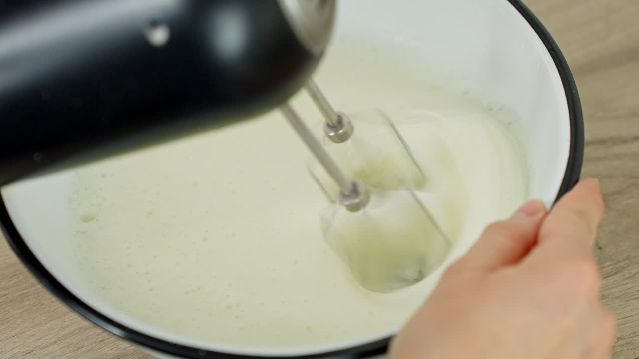 Close-up view of hand mixer beating fresh egg whites in a white bowl to stiff peaks for dessert base or meringue, shown from above in real time, making tiramisu