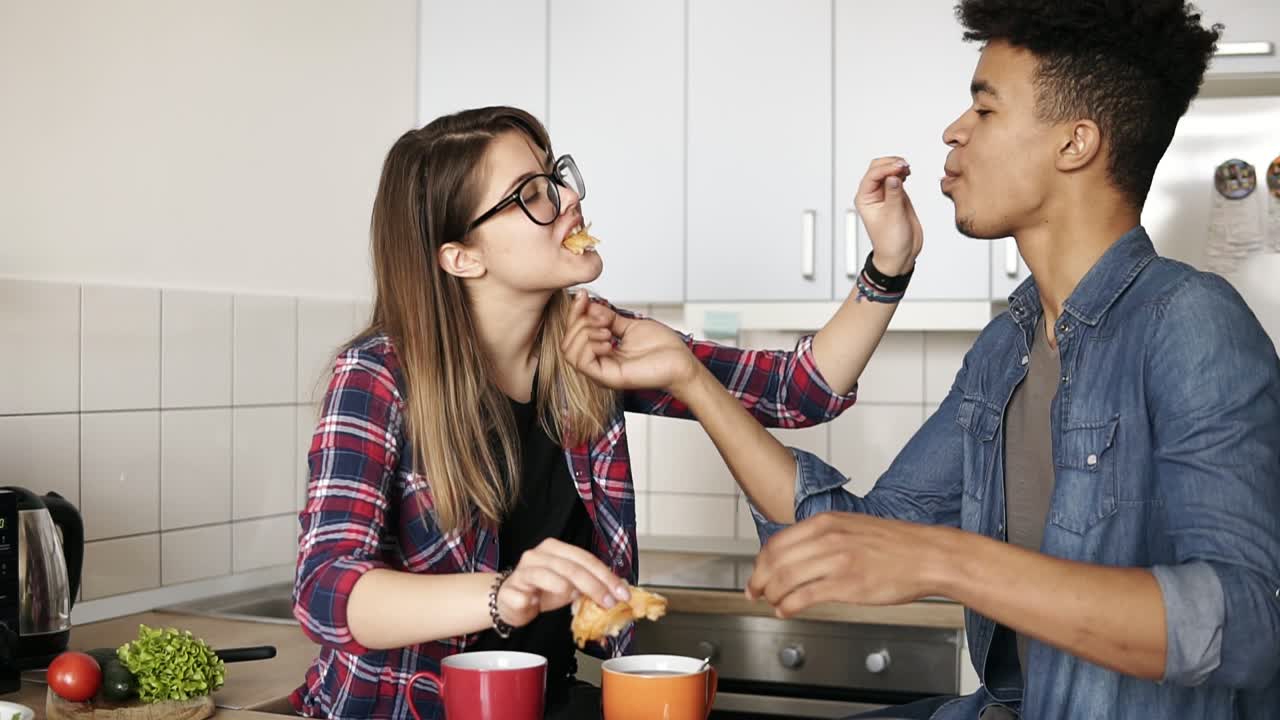 una joven pareja feliz y linda sentada en la cocina, alimentándose unos a otros con croissants, teniendo un maravilloso desayuno juntos por la mañana.