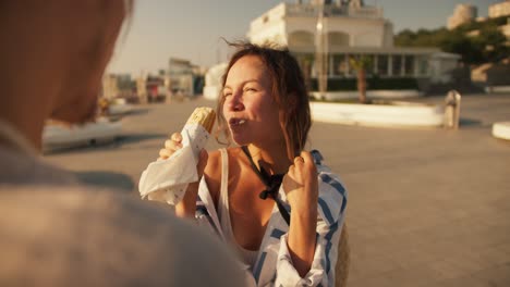 Close-up-shot-of-a-brown-haired-girl-in-a-white-blue-shirt-eating-a-hot-dog-in-the-summer-on-a-modern-beach-in-the-morning