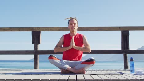 video of tranquil caucasian man with dreadlocks practicing yoga meditation sitting by sea in sun