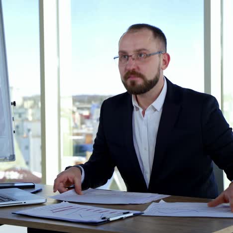 businessman in glasses at his workplace. executive man in suit sitting at desk with many papers and work in office against window background
