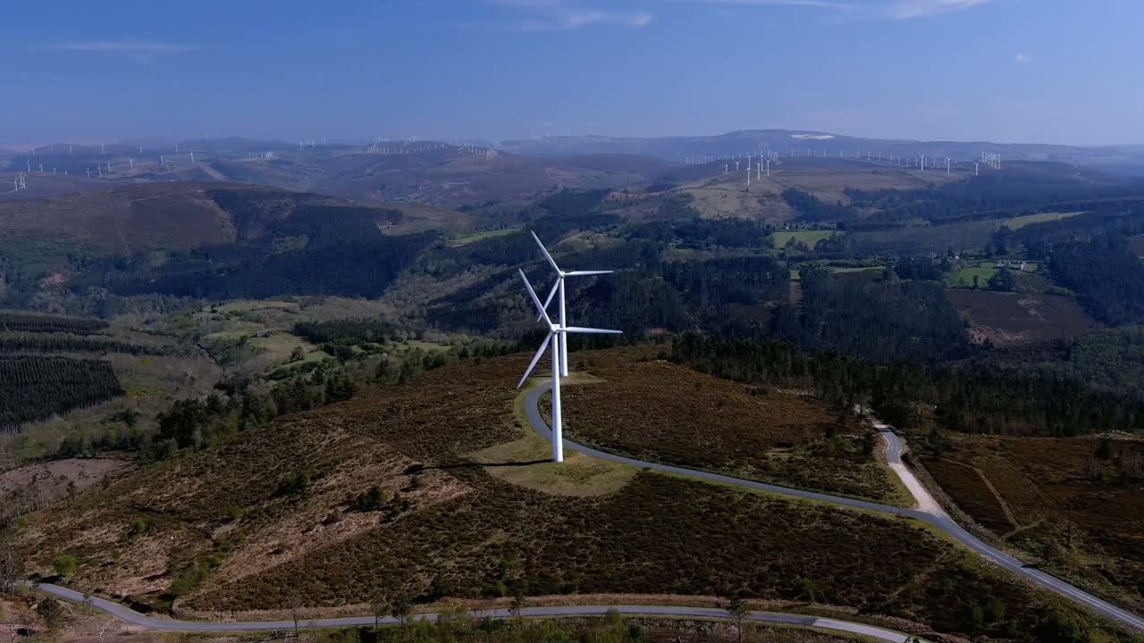 turbinas eólicas giratorias alineadas en una colina con bosques conectados por carreteras, además de turbinas eólicas al fondo en el horizonte