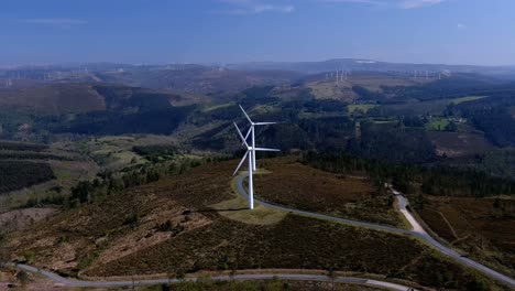 spinning wind turbines lined up on a hill with forests connected by roads, plus wind turbines in the background on the horizon