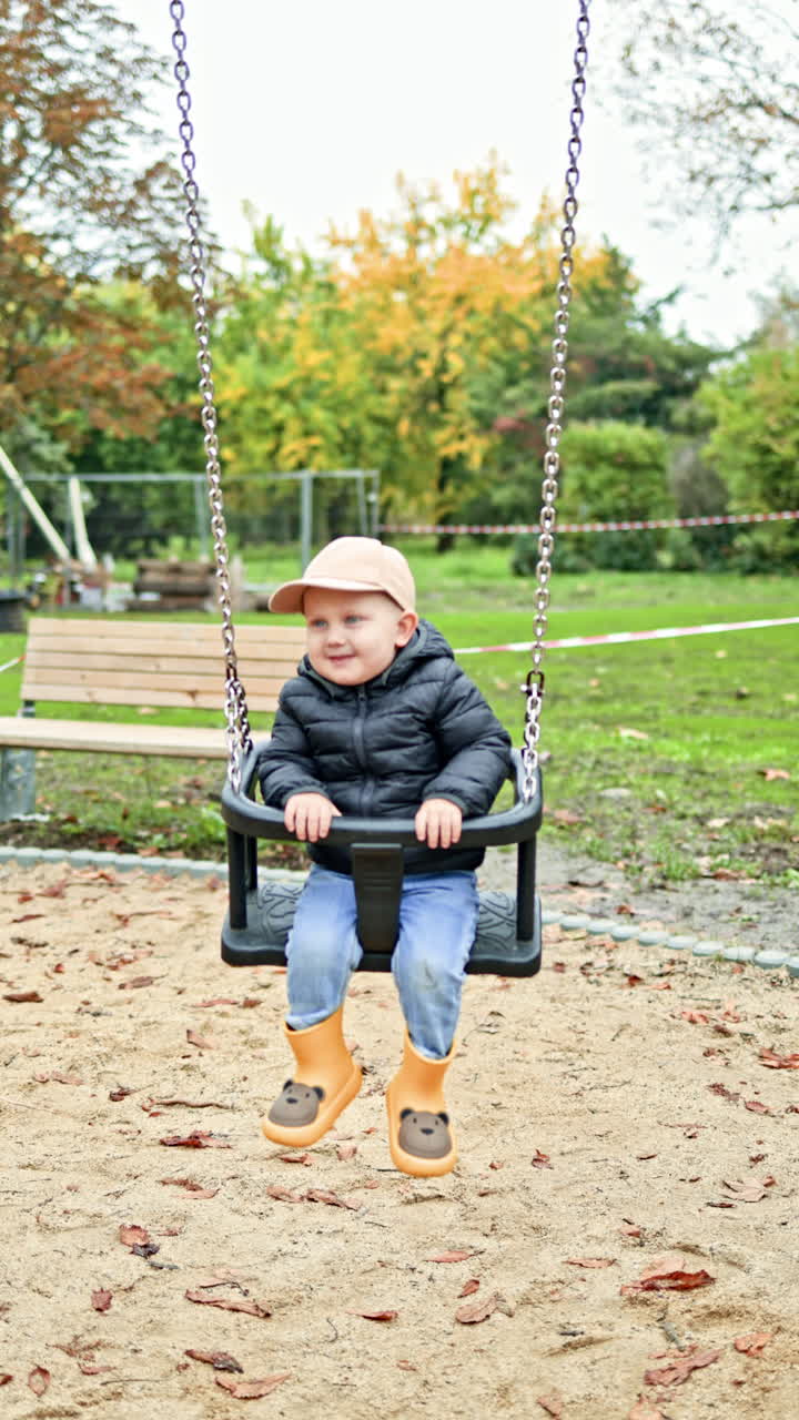 Boy 2 years old swings on a swing in a city park. Toddler swings on a playground. Child having fun and playing in the park. Children's swing on a playground