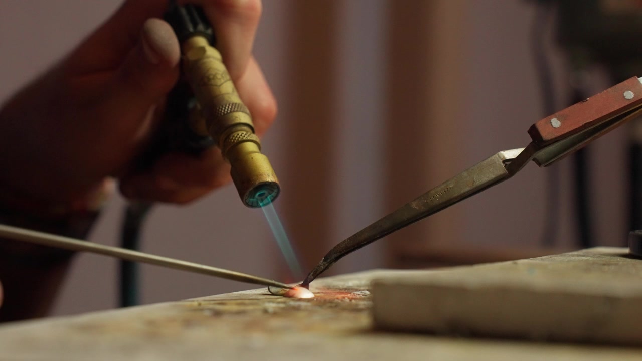 Un artigiano si concentra intensamente mentre usa una torcia per saldare componenti metallici insieme in un ambiente di officina ben illuminato, mettendo in mostra la maestria artigianale.