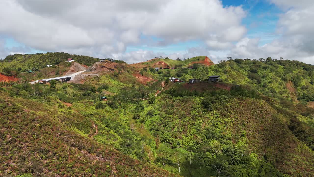 A drone view of remote mountain ridge homes surrounded by lush green hills, rolling terrain, and vibrant tropical vegetation under bright daylight with dramatic clouds