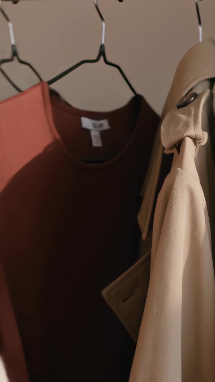 Video verticale: Mano che si allunga con maglione beige che fa scorrere un capo in maglia color ruggine su una ringhiera per controllare l'etichetta vicino a un trench.