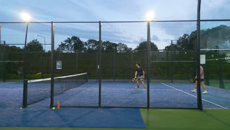 Moving drone shot from within a padel court as four players play during the evening, with artificial lighting. Dynamic yet intimate capture of sport culture