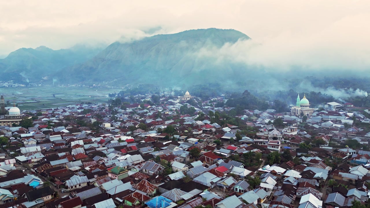Pegunungan vulkanik yang tertutup awan menjulang di atas desa Sembalun dengan masjid-masjid, rumah-rumah yang berkerumun, dan kabut yang melayang di lembah, rekaman drone udara di Lombok, Indonesia.
