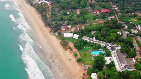 aéreo, drone inclinado disparado sobre olas y arena, en una playa paradisíaca, hacia edificios en la ciudad de trincomalee, en un día soleado, en gokanna, en la provincia oriental, de sri lanka