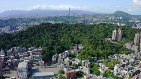 Aerial-drone-shot-of-green-growing-hills-surrounded-by-Asian-Metropolis-with-Skyscraper-buildings-during-sunny-day---Taipei-City,Taiwan