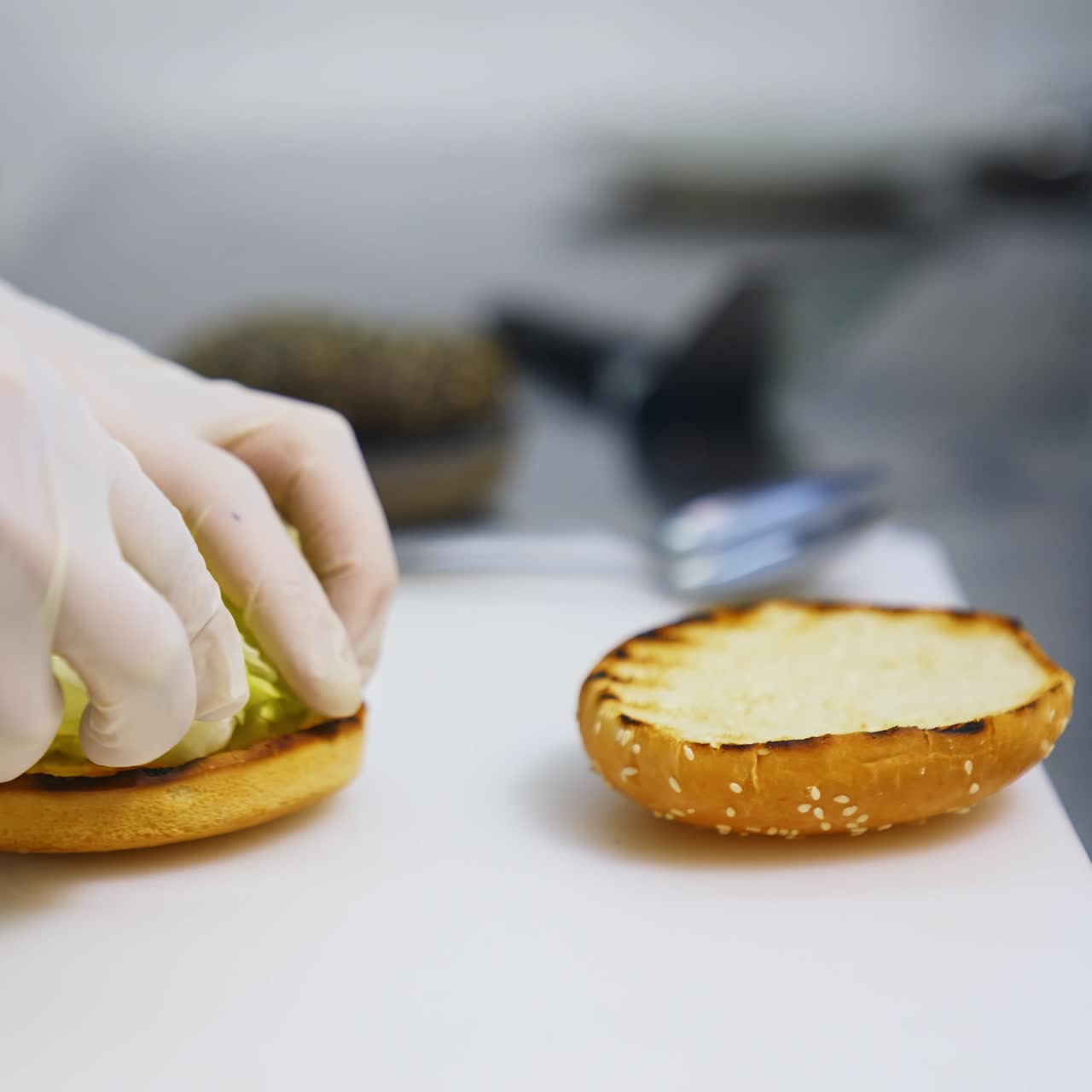 Hands in latex gloves put lettuce on top of fried bun. Making burger on white table. Fast food concept