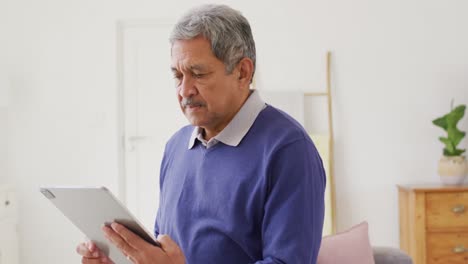 Video-of-senior-biracial-man-using-tablet-standing-in-living-room,-smiling