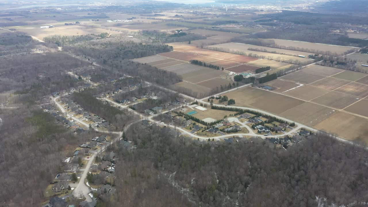 Vista aerea da drone ad alta quota di un quartiere periferico tra una foresta invernale spoglia e un vasto terreno agricolo, che mostra il contrasto tra campi rurali e sviluppo residenziale