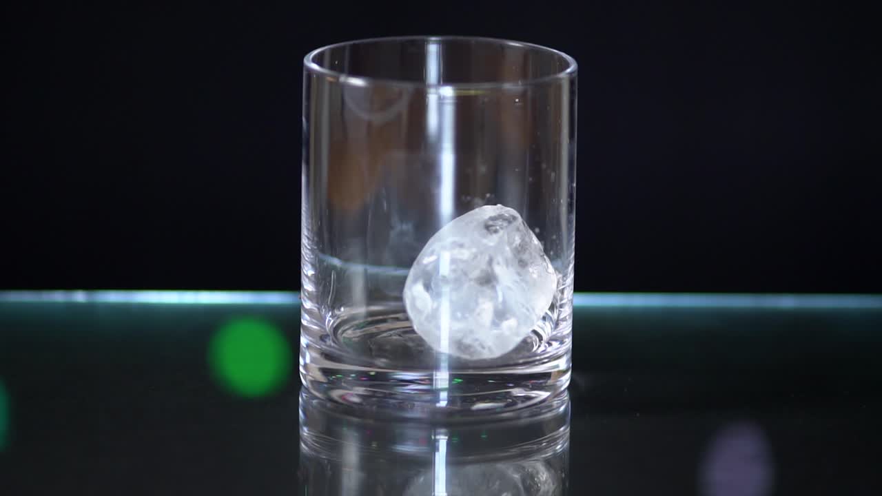 cubos de hielo gigantes cayendo en un vaso de licor sentado encima de una superficie de vidrio con un fondo negro con luces de hadas