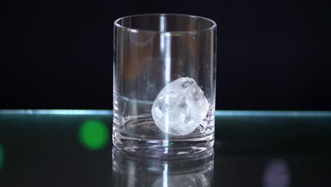 Riesige-Eiswürfel-Fallen-In-Ein-Schnapsglas,-Das-Auf-Einer-Glasoberfläche-Mit-Schwarzem-Hintergrund-Und-Lichterketten-Sitzt