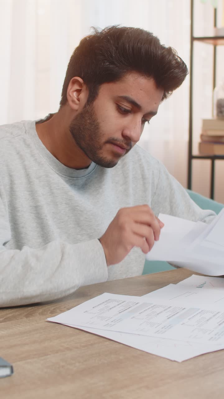 Young indian man frustrated with bills and expenses while reviewing documents at home on table