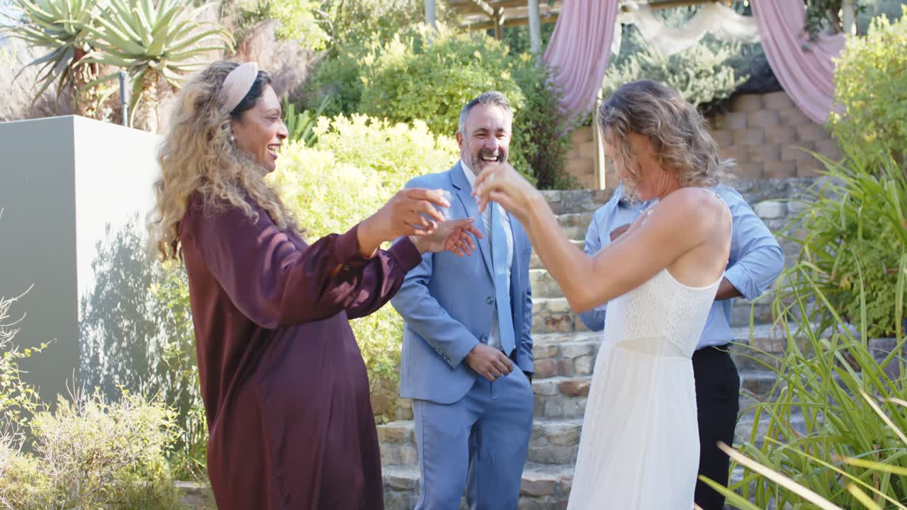 Amici di diverse etnie festeggiano su una terrazza, indossando un abito, vestiti bianchi e bordeaux, tenendosi per mano e ballando.
