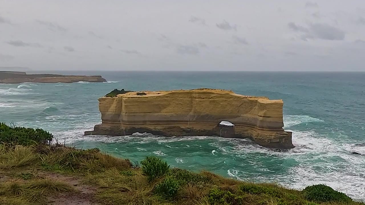 Ruwe turquoise golven beuken tegen een platte zee-boogrots voor een grasrijke kaap op een grijze, winderige dag.